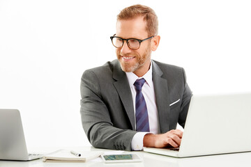 Studio shot of a smiling businessman wearing suit while sitting at desk and working on laptop. Isolated on white background.