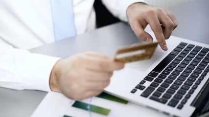 Man in white shirt holding a credit card typing information in laptop, male hands with card buying in online shop. Office manager paying using laptop and credit card