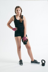 Fototapeta na wymiar Teenage girl in black sportwear and pink gloves standing with a kettlebell in her hand over the white background