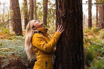 Young blonde woman in yellow coat hugging a tree in the forest and looking up to the tree top
