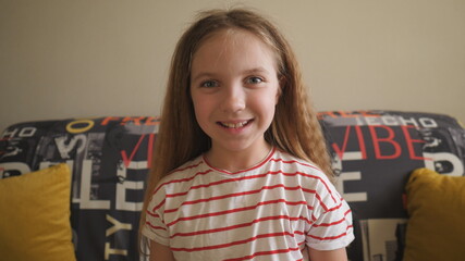 Portrait of happy blonde girl looking into camera and laughing indoor. Joyful female child sitting on the sofa at room. Close up emotions of smiling kid with glad expression on her face. Slow motion