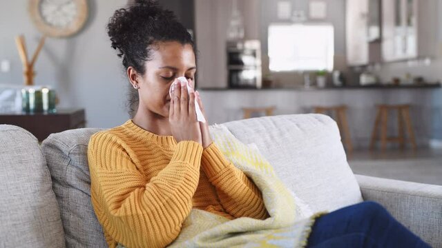 Portrait of young black woman sneezing in to tissue at home. Sick african woman wrapped in blanket sitting on sofa blowing her nose at home. Ill girl sneezing with runny nose in winter.