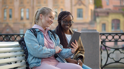 Diverse women students studying together using tablet sitting on bench outdoors
