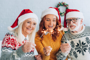 joyful senior couple with daughter in santa hats holding sparklers on christmas