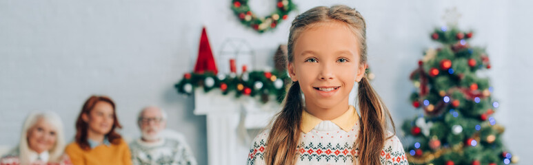 panoramic shot of happy girl looking at camera near grandparents and mother sitting on background