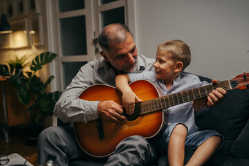 grandfather with his grandson sitting on sofa and playing guitar at home. evening scene