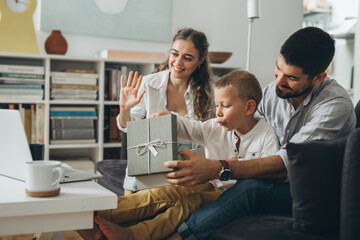 young happy family with one kid sitting on sofa and opening gifts while having video call on computer