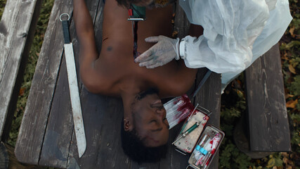 Man in coverall doing autopsy of dead afro male lying on wooden table outdoors