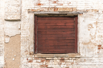 Fototapeta na wymiar Old window with brown wooden shatter on wall of abandoned building made with red bricks covered with white stucco on city street. Traditional architectural style