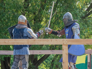 Two foot warriors in heavy medieval armor fight in the arena. Armed with swords. They are protected...