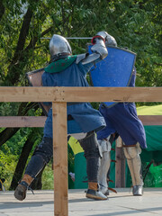 Two foot warriors in heavy medieval armor fight in the arena. Armed with swords. They are protected by iron helmets and shields. Historical reconstruction of medieval European knightly tournaments