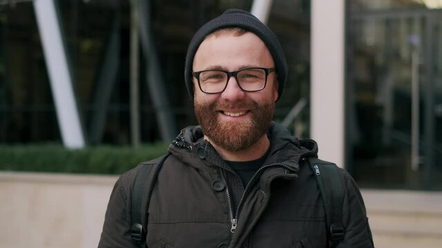 Close up view of joyful bearded guy in 30s posing while looking to camera. Portrait of handsome male person in glasseslaughing while standing at street. Concept of positive emotons.