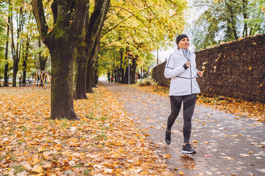 Smiling athletic woman have a jogging in the autumnal city park. Young fitness female smiling and running by the footway covered with yellow leaves. Active running people concept image.