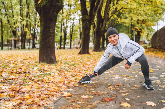 Smiling athletic woman doing workout before jogging in autumnal city park on the kids playground.Mid aged fitness female runner stretching legs while warming up. Active runners people concept image