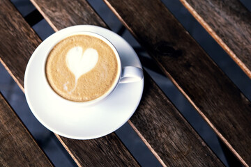 Top view of a cup of coffee on the wooden table near the street loft cafe. Trendy barista heart shape sign on the cappuccino foam or latte art. Image with copy text space.