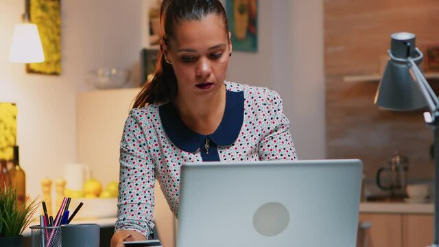 Business woman making transaction from home using credit card sitting in the kitchen late. Freelancer shopping online from home with electronic paymant on digital notebook conected to internet