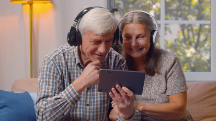 Happy elderly man and woman in headphones watching video on tablet pc at home