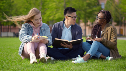 Multiethnic university students sitting and reading book on campus grass.