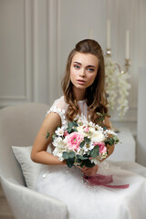 beautiful tender young bride with wedding makeup and long curly hair holding flower bouquet and sitting in armchair in white studio interior