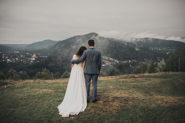 Fototapeta na wymiar bride in a wedding dress stands her back groom in a classic suit looking at the forest in the mountains in the fog