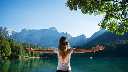 Young woman with long brown hair standing by a beautiful green lake with her arms spread widely