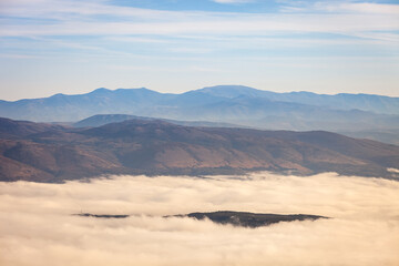 Hill raising above the thick fog covered by forest and distant, impressive horizon mountains - 386728796