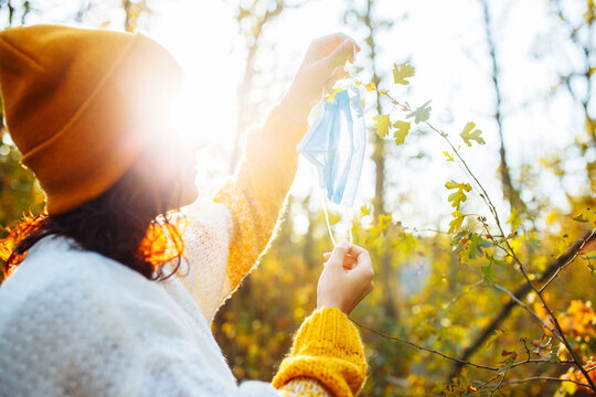 Young Woman Wearing An Orange Hat And Sweater Hangs Out The Medical Sterile Mask On The Tree In The Autumn Forest. Female Breathes Fresh Air In Fall Park During Coronavirus Covid19 Pandemic. Health.