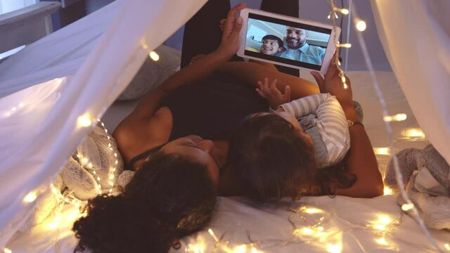 Mother and cute girl using digital tablet while lying in kid tent doing a video call with father and son. Indian family in online conversation with each other during quarantine and social distancing.
