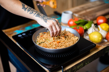 Mujer tatuada cocina comida oriental sobre sartén en placa de vitroceramica y verduras. Mujer sazonando la comida.
