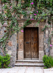 A very old door with a cement frame and a creeper with pink flowers over a stone wall