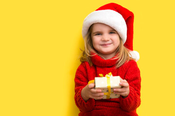 Child in a red sweater and a Santa hat  with Christmas gift box against yellow background. Christmas shopping.  Copy space. 