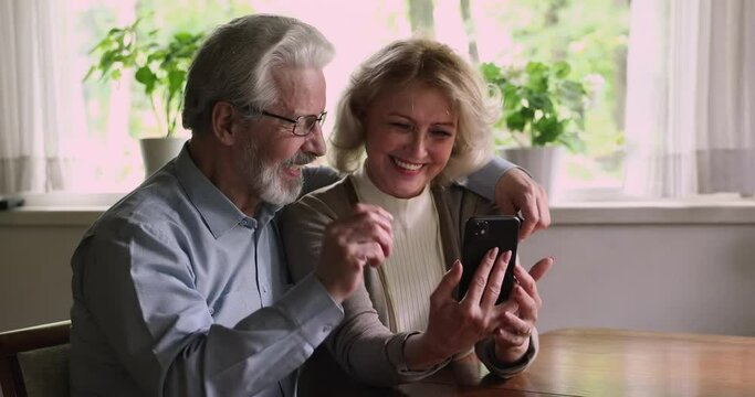 Middle-aged wife and husband sitting resting at home using mobile phone, talking having fun on internet, watch videos and photos online, discuss new software. Comfort modern technology usage concept