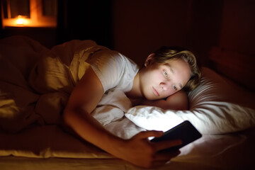 Teenager boy chatting by phone, writing in blog or reading social networks instead sleeping. Young...