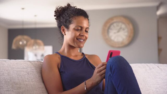 Cheerful young african woman using smartphone while sitting on couch. Black smiling woman using app on cellphone at home. Beautiful girl relaxing while chatting on mobile phone and looking at camera. 
