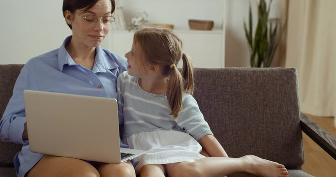 Happy mum and cute kid daughter having fun sitting at sofa at home talking using learning laptop, smiling mother teach little child girl online, make webcam video call looking at computer screen
