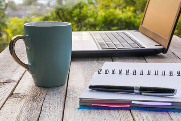 Laptop, notebook with pen and cup of coffee on a wooden table. Workplace outdoors