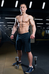 Fototapeta na wymiar A shirtless bodybuilder in shorts is posing full length with two dumbbells on the floor in a gym. A caucasian men's physique athlete is relaxing after an exercise.