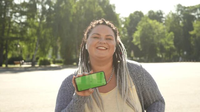 Beautiful stout girl with chromakey on her smartphone advertising mobile app. Commercial, copy space concept. Young pretty fat woman with dreads showing green screen phone to camera and dancing.