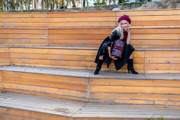 A happy girl sits on a summer theater bench, made of wood, in a burgundy coat and biret, an adult with tender lips, in the fall against a background of blue clouds.