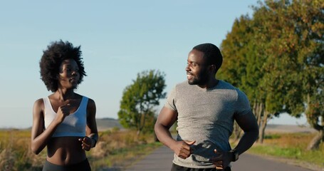 Cheerful African American young sporty man and woman joggers running on road early in the morning in countryside and talking. Communication. Happy sportswoman and sportsman jogging in nature.