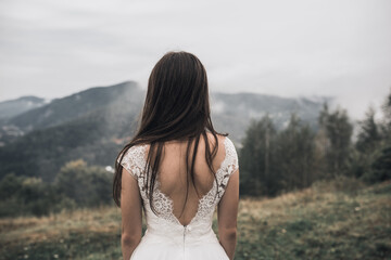 A thin young brunette with long hair, a bride in a white wedding dress with open back, stands in the middle of a green meadow. Against the background of large mountains covered with trees in the fog.