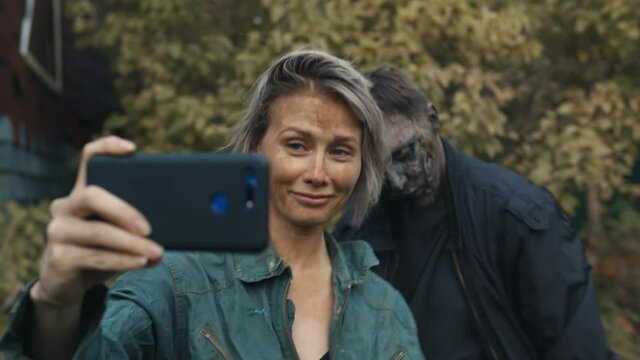 Young woman taking selfie with zombie on smartphone outdoors