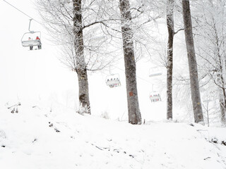 A chair lift with people on a winter foggy snowy day drives past the trees growing on a hillock