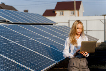 Investor woman stand with laptop near blue solar panels row on the ground Girl weared formal white shirt. Free electricity for home. Sustainability of planet. Green energy.