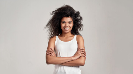 Portrait of young african american woman with curly hair wearing white shirt smiling at camera while standing with arms crossed isolated over grey background