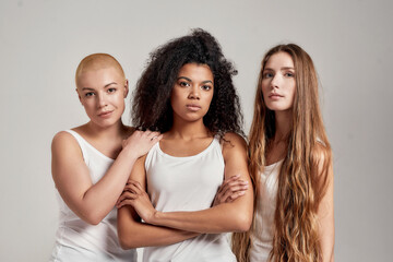 Portrait of three young diverse women wearing white shirts looking at camera while posing together isolated over grey background