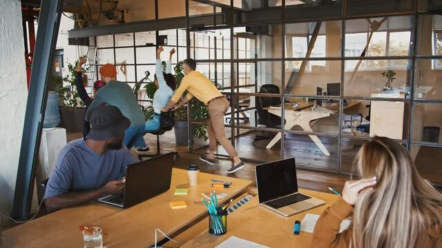 Diverse managers watching their colleagues competing in office chair race at work, slow motion