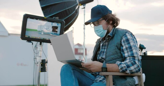 Portrait of Caucasian male filmmaker in mask and glasses sitting into director chair and looking into screen on set while shooting video outdoors. Man with laptop on backstage. Cinematography concept