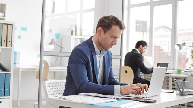 Slow motion of male Caucasian business person typing on keyboard, closing laptop cover, rubbing eyes and walking away, colleague working in background, man walking by camera