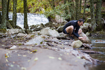 Hombre cogiendo agua de un rio natural en el bosque en una garganta de piedras en medio de un valle con una cascada de agua de fondo en un entorno natural.